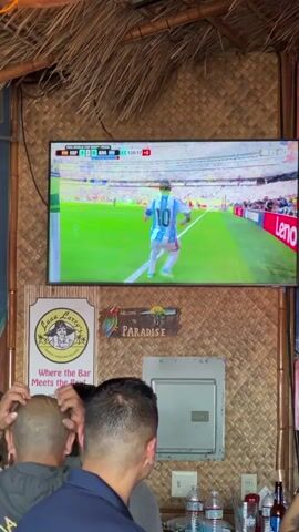 The video shows a group of men watching a soccer game on a television screen. The game appears to be in overtime, as indicated by the timer on the screen. The men are standing and looking intently at the television, with some of them appearing to be cheering or reacting to the game. The television is mounted on a wall covered in a woven material, possibly bamboo. Below the television are two signs, one with the text "Lazy Larry's Where the Bar Meets the Rest" and another with the word "PARADISE" and a picture of a parrot. There are bottles of water and other drinks on a counter in front of the men. The overall environment suggests a bar or casual gathering place.