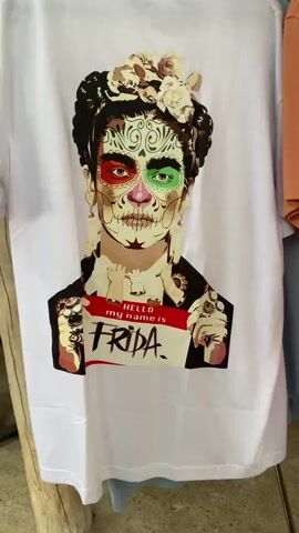 A close-up shot shows a white t-shirt hanging, featuring a print of Frida Kahlo with sugar skull makeup. The left eye is painted red and the right eye is green. She is wearing earrings, rings, and a floral headpiece. The t-shirt design also includes a name tag that reads, "Hello, my name is Frida." A wooden post is visible in the background to the left. Another item of clothing, peach-colored, is to the right. The camera slowly zooms in closer on the image on the t-shirt.