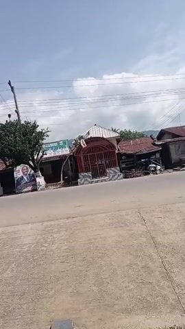 The video pans across a street scene with several buildings on the right side of the road. The buildings appear to be small shops or houses with corrugated metal roofs. There is a red metal gate in front of one of the structures. A large poster with a picture of a man is visible on the left side of the road. Overhead power lines are strung across the scene. The sky is partly cloudy.