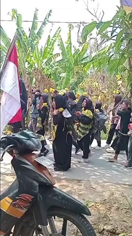 The video shows a group of people, primarily women and girls, walking along a paved path outdoors. Many of them are wearing black outfits with yellow stripes, some resembling bee costumes. One person in the group is carrying a flag that appears to be the Indonesian flag, red and white. Some individuals are also wearing headscarves. The environment includes lush green banana trees and other vegetation, suggesting a tropical or subtropical setting. In the foreground, the front of a motorcycle is visible. The people are moving from left to right across the frame. Some are interacting, talking, and looking around. The lighting indicates it is daytime with bright sunlight.