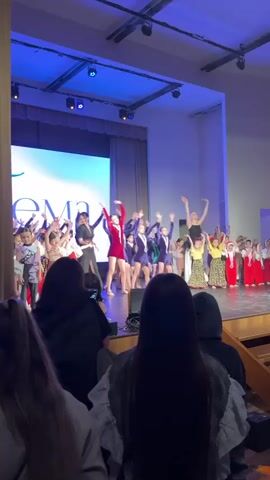 The video shows a group of children performing on stage in front of an audience. The stage is brightly lit with blue lights, and a large screen displays white text. The children are dressed in various costumes, including red dresses, navy blue suits, yellow dresses, and red outfits with white trim. They are performing a dance routine with their arms raised. Several adults are standing with the children, possibly as instructors or chaperones. The audience members are seated in rows and watching the performance, with some holding up their phones to record.