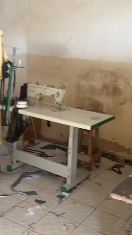 The video shows a sewing machine on a white table in a room with off-white walls. The sewing machine is labeled "TWO LION INDUSTRIAL SEWING MACHINE CO.LTD." There are scraps of fabric on the tiled floor, along with a black trash bag hanging off the side of the table. Various sewing tools and accessories are on a shelf above the table, including measuring tapes and spools of thread. A blender and a yellow mug are partially visible in the lower left corner of the frame. The walls appear worn, with some discoloration and possible water damage.