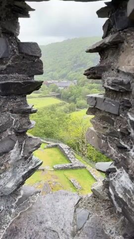 The camera peeks through a narrow stone opening, revealing a lush green landscape on an overcast day. In the foreground, stone foundations sit atop a grassy hill, surrounded by a wire fence. Beyond this, a river winds through fields and trees towards a lake nestled against a forested mountain. A road with sparse traffic traces the riverbank. The stone opening frames the scene, emphasizing the contrast between the rough, aged rock and the vibrant natural beauty of the landscape.