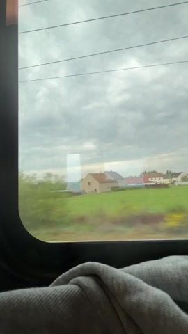 The video shows a view out of a train window. The train is moving through a rural area with green fields and scattered houses. The sky is overcast. There is a gray blanket or sweater visible in the foreground.