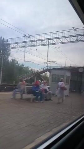 The video shows a view from a train window. The train is moving, as indicated by the blur and the passing scenery. The first few seconds show the interior of a train station, with a platform, a covered walkway, and a person waiting on the platform. The train then passes a series of cargo containers on a parallel track. The environment appears to be semi-urban or rural, with trees and power lines visible. The sky is overcast.