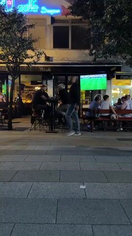 The video pans from left to right across an outdoor seating area of a cafe or restaurant at night. A neon sign that reads "Café" with some unreadable text underneath is visible at the top left. Several people are seated at tables and bar stools, some appearing to be dining or drinking. A large television screen displaying a soccer game is mounted on the wall of the establishment. A tree with dark green foliage stands in front of the television. To the right, a yellow food kiosk with an awning is set up. Another tree with similar foliage is positioned between the cafe and the kiosk. A few more people are visible seated outside the kiosk. The ground is paved with interlocking grey tiles. The overall atmosphere appears to be casual and social, with people enjoying themselves outdoors in the evening.
