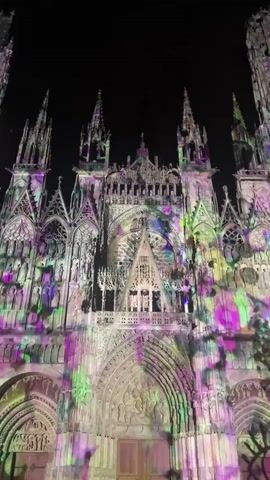 A low-angle, static shot shows the ornate facade of a Gothic cathedral at night. The cathedral's light-colored stone is entirely covered by a dynamic light projection featuring swirling and splattering patterns in various colors, including purple, green, yellow, and blue. These colorful patterns move and change across the cathedral's intricate architectural details, including its multiple spires, statues, large rose window, and grand arched entrance. The sky behind the cathedral is dark black.