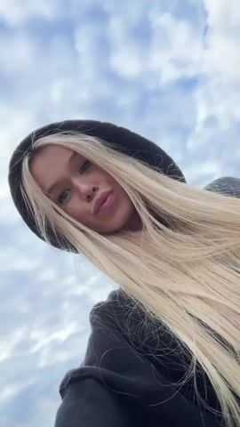 The video is a selfie-style recording of a young woman with long, blonde hair and a black hoodie. She is outdoors with a cloudy sky and some greenery in the background. The woman smiles at the camera and makes playful facial expressions. The video appears to be shot on a smartphone and is oriented vertically.
