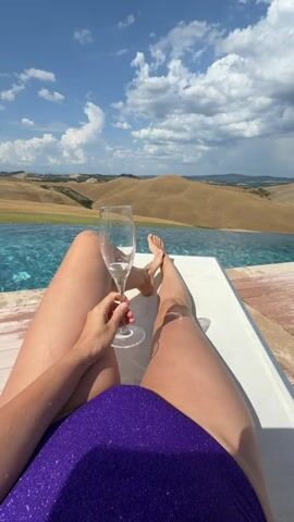 The media file features a first-person perspective shot of a person relaxing on a lounge chair next to an infinity pool. The person is wearing a sparkling purple swimsuit and holding a champagne flute. Their legs and feet are visible, along with their arm and hand holding the glass. The background showcases a picturesque landscape of rolling hills, characteristic of the Tuscan countryside in Italy. The sky is blue with scattered white clouds, indicating fair weather. The time of day appears to be daytime, likely late morning or early afternoon, given the bright sunlight casting shadows. A small, light-colored dog with curly fur is seen in the middle ground, near the edge of the pool, looking towards the camera. The overall scene evokes a sense of leisure, relaxation, and enjoyment of a beautiful day in a scenic location. There is no visible text in the media file.
