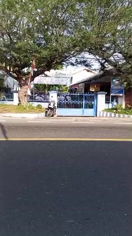 A person wearing a brown uniform and a cap rides a green scooter down a paved road. The scooter is moving from left to right in the frame. The rider is looking forward. In the background, there is a building with a blue gate and a sign that reads "NEGERI 2 PANCUR BATU". A black motorcycle is parked on the left side of the road, near the gate. Several people are visible standing behind the gate. The environment appears to be outdoors on a sunny day. The road has a yellow line painted down the center.