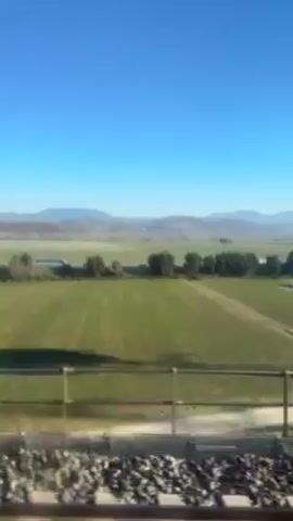 The video shows a view from a moving train. The train tracks and gravel are visible at the bottom of the frame. A low fence runs along the side of the tracks. Beyond the fence is a large, flat field of green grass. In the distance are low mountains under a clear, bright blue sky. There are a few trees and small structures scattered across the field. The camera is shaking slightly due to the movement of the train.