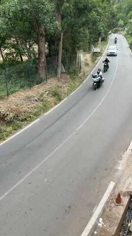 A group of motorcyclists rides along a paved road in Asgaour, Morocco. The video opens with a shot from an elevated position, looking down at the road. Two people on a grey touring motorcycle, both wearing helmets, pass by the camera. Behind them, another rider on a black motorcycle is visible. As the camera pans to follow the motorcycles, more members of the group come into view, indicating a larger convoy of at least five to six motorcycles.

The road is bordered by a mix of green trees, dry brush, and a chain-link fence. On the right side of the road, several cars are parked near buildings with distinct Moroccan architecture, including a white structure and a pinkish-orange building with blue doors. The background reveals a mountainous landscape, covered with sparse vegetation, suggesting a valley or mountain pass. Power poles line the road, and the overall impression is of a scenic, rural setting. The time is daytime, and the weather appears clear, with bright natural light illuminating the scene.