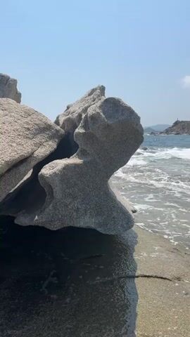 The video opens with a close-up shot of a large, weathered rock formation on a beach. The rock is gray and has an unusual, sculpted shape. Gentle waves lap at the base of the rock, and the water appears clear. The camera then pans to the right, revealing the ocean, a rocky coastline, and a distant hill covered in green vegetation. The sky is blue with a few scattered white clouds. The waves continue to roll in, creating white foam as they reach the shore. In the distance, a small lighthouse or tower is visible on the headland. The video then shifts to a wider shot of the beach and the sea. More waves are breaking, and the water is a beautiful turquoise color. In the background, several people can be seen on the beach, some sitting under umbrellas and others swimming in the water. Sailboats are visible on the horizon, adding to the scenic view. The camera continues to pan, showing the coastline stretching out, with more hills and mountains in the distance. The clear water reveals rocks and sand beneath the surface. The video concludes with a final wide shot of the idyllic beach scene, bathed in sunlight.