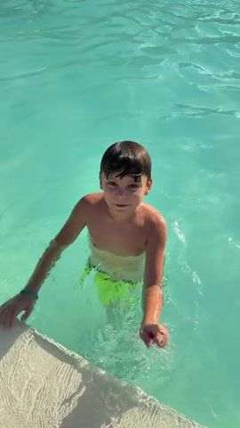 A young boy with short dark hair and wearing green swim trunks is standing in a swimming pool. The water is a clear turquoise color. The boy is holding onto the edge of the pool with both hands. He is looking towards the camera and smiling. The pool edge is textured and appears to be made of concrete or stone. The sun is shining, casting reflections on the water.