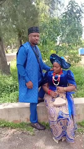 A man and a woman are posing for a video outdoors. The man is standing on the left, wearing a blue kaftan and pants with a patterned design on the chest and sleeves. He has a black patterned cap on his head and is wearing dark shoes. The woman is sitting on a low concrete wall on the right, wearing a blue velvet top with puffed sleeves and a blue and gold patterned skirt. She has a blue headwrap, a layered red necklace, and is holding a beige purse. The man is looking at the camera and the woman is looking down at her purse. The background consists of trees, bushes, and a gray paved area. The video shows the man approaching the woman, placing his hand on her arm, and then walking away from the camera, turning his back to the woman. The woman remains seated.