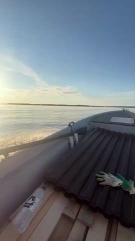 The video shows the view from a moving boat on a large body of water. The sun is setting, casting a golden light on the water and the sky. The boat is an inflatable vessel with some cargo on board, including what appears to be roofing material, a plastic bottle of water, and a pair of work gloves. The wake of the boat is visible, indicating forward motion. The horizon is distant and hazy. The overall impression is one of calm and tranquility, with a sense of adventure and purpose implied by the cargo.