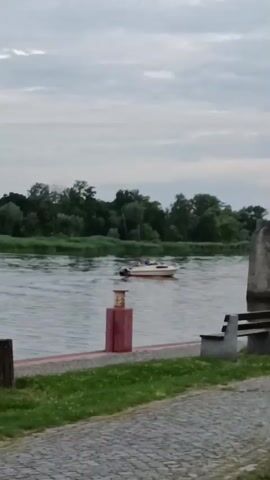 A video shows a motorboat moving on a river. On the riverbank, there is a red bollard with a light, a bench, and a stone structure. Trees are visible in the background. The sky is cloudy.