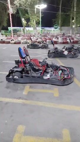 The video pans across a go-kart track at night. Several go-karts are parked in a pit area, some with their engines exposed. The track itself is lined with tires stacked in a barrier. Overhead lights illuminate the scene, and trees are visible in the background. There are no people visible on the track or in the immediate vicinity. The words "START" are visible on a sign in the background, and the word "STOP" is painted on the track surface in the foreground.