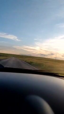 The video begins with a view from inside a car, looking out through the windshield at a two-lane asphalt road winding through a vast, grassy landscape under a blue sky with scattered clouds. The sun is low on the horizon, casting a warm glow and creating a slight haze. The landscape appears to be a rural, open field with sparse vegetation and a few utility poles visible along the roadside. The car's dashboard is visible in the foreground, obscuring the bottom portion of the frame. The camera is stationary, capturing the scene as if the car is moving forward. The audio consists of faint music and some indistinct speech, possibly from a radio or conversation within the car. The overall impression is a serene drive through a countryside at dusk.