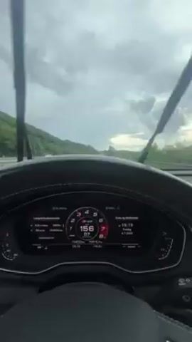 Certainly! Here's a detailed description of the video:

**Scene:** The video presents a driver's perspective from inside a car moving along a highway.

**Environment:**

*   The exterior environment is characterized by an overcast sky with visible cloud formations.
*   The highway is flanked by lush green hills or slopes.
*   The road appears wet, possibly from recent or ongoing rain.

**Objects:**

*   The primary focus is the car's dashboard, featuring a digital speedometer displaying the vehicle's speed, which varies around 155-157 km/h.
*   Other information visible on the dashboard includes trip data, time (19:19), and date (4.7.2023).
*   The steering wheel is visible, with control buttons and trim details.
*   Other vehicles can be seen moving on the highway in the distance.

**Actions:**

*   The car is in motion, moving along the highway.
*   The driver is maintaining a relatively high speed.
*   Minor fluctuations in speed are visible on the speedometer.

**Interactions:**

*   The video is primarily documenting the driving experience.

**Additional Notes:**

*   The perspective is static, focusing on the view through the windshield.
*   The overall tone suggests a regular driving scenario, possibly during a road trip.