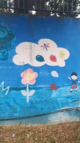 The video shows a mural painted on a blue wall. The mural depicts a stylized landscape with clouds, a rainbow, flowers, a snail, a dragonfly, balloons, and a child. The style of the painting is childlike and colorful. The wall appears to be outdoors, with some greenery and dry leaves in the foreground. A metal fence is visible at the top of the wall. The video pans slowly across the mural, showing different parts of the artwork.