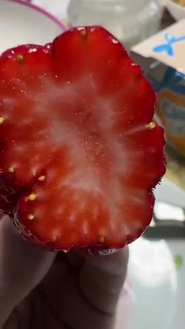 Here's a description of the video:
The video shows a person preparing strawberries in a home setting. The camera focuses on a hand holding a half of a strawberry, revealing the red flesh and visible seeds. The camera then pans out to show a table laden with items including a jar of white powder, a box of cookies labeled "Сладкие гостинцы" (Sweet Treats), and a glass of dark liquid. Another person wearing a green jacket is shown picking up strawberries from a pink bowl. The bowl contains several bright red strawberries with green stems attached.