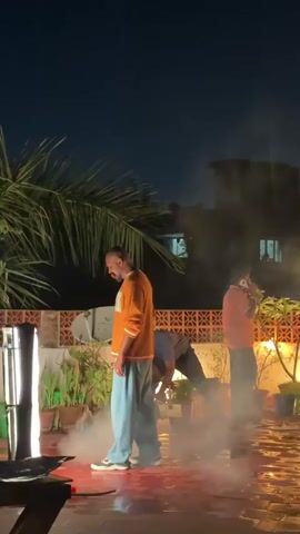 The video captures a nighttime outdoor scene, likely a rooftop or terrace, in an urban setting. The area features a brick wall, potted plants, a large palm tree, and a satellite dish.

The central subject is a bald man wearing an orange long-sleeved shirt, light blue baggy pants, and white sneakers. He has a dramatic red marking, possibly artificial blood, on his face near his mouth. He is performing an action, initially standing and then transitioning into a low, crouching pose, as if in distress or intense focus. Assisting him is another man in a dark shirt, kneeling behind him. A third man, wearing a pinkish-red shirt, stands further back on the right, talking on a phone, presumably observing or directing the scene.

The ground is covered in a dense layer of low-lying fog or smoke, with visible patches of a reflective, wet surface, and some reddish liquid or staining, possibly fake blood, particularly around the central figure. A tall, rectangular artificial light source illuminates the scene from the left. The word "Action" is audible at the beginning, indicating that a film or photography shoot is in progress. The overall ambiance is dramatic and intense, characteristic of a production set.