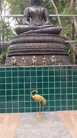 A static medium shot shows a large bronze Buddha statue seated on a tiered base. The statue has multiple faces stacked on its head. In front of the statue, on a platform covered in green tiles, is a small golden crane sculpture. The background consists of trees and foliage, suggesting an outdoor setting. Metal poles are visible to the right of the statue.