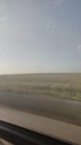 This video shows a view from inside a vehicle looking out a window. The window frame is visible in the foreground on the left and bottom of the frame. The sun is shining brightly from the upper left, casting reflections on the window. Outside, there is a wide, flat, grassy field stretching to the horizon. A dirt road or path is visible in front of the field. The sky is mostly clear with a few faint clouds. The scene appears to be a rural or open landscape. The video is a short, still shot of this view.