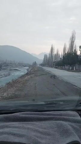 The video shows a view from inside a car driving along a road next to a river. The road is paved and stretches into the distance, bordered by trees on one side and the river on the other. A bridge crosses the river further down the road. Mountains are visible in the background, shrouded in mist or fog. The sky is overcast. The car's dashboard is visible in the foreground, covered with a grey cloth. The scene appears calm and quiet.