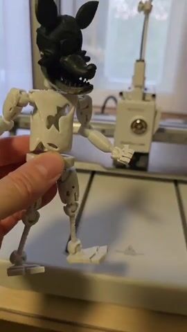 A person's hand is holding a 3D printed, articulated toy robot that resembles an animatronic character. The robot is mostly white with black accents on its head, which features a mask with a menacing expression and sharp teeth. The robot has visible joints at its limbs and torso, indicating it is designed for posing. One of the robot's feet is partially detached, with pieces lying on the surface beneath it. In the background, a white 3D printer with the "Bambu Lab" logo is visible, with its print head positioned over the print bed. The overall environment appears to be a workshop or indoor space, with a window in the background showing daylight and greenery outside. The video shows the robot being manipulated by the hand as the camera pans slightly.