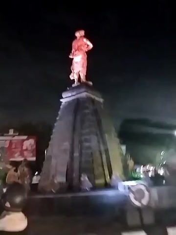 The video begins by showing a large statue illuminated at night. The statue depicts a person standing on top of a multi-tiered, pyramidal base. The statue itself is lit in a reddish-orange hue. As the camera pans, the base of the statue is revealed to have smaller figures and structures around it, also lit with colored lights, primarily pink and green. The scene appears to be outdoors, with trees and dark sky visible in the background. The camera then moves to show a street scene at night. Vehicles, including motorcycles and cars, are present and appear to be in traffic. People are seen on motorcycles, wearing helmets. Another, smaller statue or figure is visible in the distance, also illuminated, appearing to be on a pole or elevated platform. The overall impression is a busy urban environment at night, with a prominent illuminated statue as a central feature. The video is shaky and the lighting is dim in parts, suggesting it was filmed handheld at night.