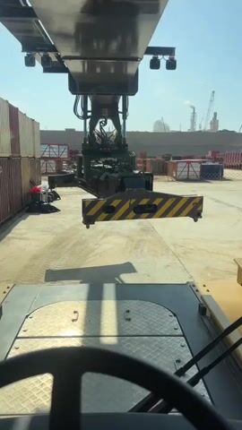The video is filmed from the perspective of someone inside a large piece of industrial machinery, likely a container handler or crane. In the foreground, a steering wheel is visible, along with parts of the vehicle's dashboard. The main focus is the massive structure of the machinery extending forward and upward. A yellow and black striped attachment, possibly a spreader or grapple, hangs suspended in the air, indicating it is in the process of moving or has just finished moving something. In the background, stacks of shipping containers are lined up on either side, suggesting a port or industrial yard. Further in the distance, industrial buildings with smokestacks and other large structures like cranes are visible under a clear, sunny sky. The overall environment appears to be a busy, operational industrial site.