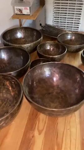 The video shows a collection of bronze singing bowls arranged on a wooden floor. The bowls vary in size, with some appearing larger and others smaller. They have a hammered texture and a dark, metallic finish. In the background, there are shelves with some objects on them, including a plastic container with labels and what appears to be a bundle of sticks or rods. There is also a white radiator or vent-like structure visible. The lighting is somewhat dim, creating reflections on the surface of the bowls. No people are visible in the video, and the focus is entirely on the arrangement of the singing bowls.
