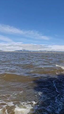 The media file shows a view from a boat moving across a body of water. The water is choppy with whitecaps, suggesting a windy or rough condition. In the distance, a shoreline is visible with some snow-capped mountains or hills. The sky is a clear, bright blue with scattered white clouds. The overall impression is of a scenic, possibly remote natural landscape. The time of day appears to be daytime, likely late morning or early afternoon, given the bright sunlight. There are no people or identifiable objects in the frame, apart from the implied boat from which the video is recorded. The location context provided is Anadyr, Russia, which aligns with a potentially cold, mountainous, and coastal environment. No text is visible in the media file.
