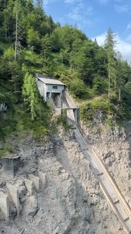 The video shows a steep, rocky cliff face with a body of turquoise water at its base. On the cliff, a concrete structure with a slanted track or pathway runs down from a building situated higher up. The building appears to be made of concrete and is partially overgrown with greenery. Trees and vegetation cover the upper part of the cliff and the surrounding hills. The water is calm, and a few buoys are visible in the distance. The sky is blue with scattered white clouds. The scene suggests a dam or reservoir in a mountainous, forested area.