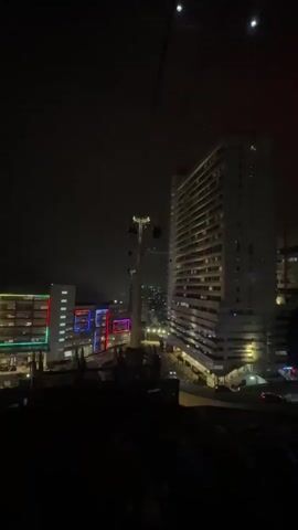 The video captures a nighttime view of a city scene from what appears to be a cable car or gondola lift. In the foreground, the dark silhouette of the cable car structure is visible. Further out, a tall, white high-rise building stands prominently, its windows faintly illuminated. To the left of the high-rise is a shorter building with colorful, square-shaped lighting in green, red, and purple. Streetlights illuminate the roads and walkways below. The background is dark, with a few distant lights suggesting the presence of other buildings or city infrastructure. The overall scene is somewhat dark, with the main light sources being the building illuminations and streetlights.