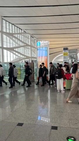 A group of people are walking through a modern airport terminal. They are walking from left to right across the frame. Most of the people are wearing casual clothing and carrying luggage, such as backpacks and suitcases. There are signs in the background with Chinese characters and English text, indicating that this is likely an international airport. The floor is polished and reflective, and the ceiling has a distinctive ribbed design. The overall impression is one of a busy travel hub.