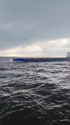 A long blue cargo ship sails across dark, choppy water under a cloudy sky. The ship is mostly empty, with a visible railing running along its deck. The camera appears to be on another vessel, moving parallel to the cargo ship.