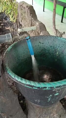 Water is flowing from a blue hose into a large black plastic tub. The tub appears to be partially filled with dark-colored water and some debris. The hose is attached to a faucet that is visible in the background, along with a pipe. To the left of the faucet, there are some green plants. Behind the tub, there is a textured, light-colored structure that resembles a rock or a decorative element with some writing on it. The ground around the tub is dirt and gravel. The scene appears to be outdoors, possibly in a garden or courtyard, with a green wall visible in the far background. The overall lighting suggests it is daytime.