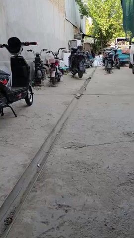 The video opens with a view of a dusty, outdoor area with a concrete floor. Several parked electric scooters are visible. The camera pans to the right, revealing a white SUV and more scooters. The focus then shifts to a close-up of an electric scooter, showing its thick tires and suspension. The scene transitions to a pile of discarded electronic components, including circuit boards, wires, and a toolbox, suggesting a repair or recycling area. The video ends with a shot of the messy collection of electronics.