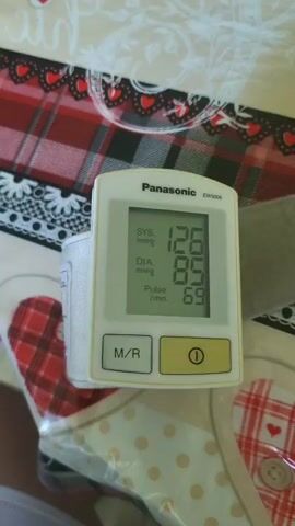 A Panasonic EW3006 blood pressure monitor displays a systolic reading of 126 mmHg, a diastolic reading of 85 mmHg, and a pulse of 69 beats per minute. The device has two buttons: M/R and a yellow button with a power symbol. The monitor is placed on a patterned tablecloth with red and white plaid and white polka dots.