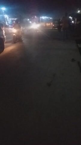 The video captures a night scene from a moving perspective, likely from a vehicle such as a motorcycle or scooter, given the view of handlebars and the sound of an engine and music. The camera's orientation shifts from an initially tilted vertical to a clear vertical (portrait) view, and then briefly to a horizontal (landscape) orientation before returning to vertical.

On the left side of the frame, numerous artificial lights are visible. These lights, predominantly bright white and blue, with some occasional orange or yellow hues, appear as streaks and blurred spots due to motion. They likely represent streetlights, lights from other vehicles, or buildings lining the street. These lights create a bright, dynamic streak along the left edge of the video.

The right side of the frame is dominated by a large, dark, and mostly featureless wall or building. Its texture is somewhat visible, suggesting concrete or similar material, but the darkness prevents clear identification.

In the foreground, particularly at the beginning of the video, parts of a vehicle's handlebars and the top of a front wheel or dashboard are dimly visible, reinforcing the first-person perspective from a moving vehicle. The overall lighting is dim, characteristic of nighttime conditions, with the primary illumination coming from the artificial light sources themselves. The scene conveys the experience of traveling at night, with the surrounding environment largely obscured except for the prominent lights.