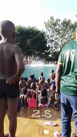 The video shows a group of children in a swimming pool. They appear to be sitting or standing in the shallow end of the pool. Several adults are standing on the pool deck. The pool is surrounded by trees and other people can be seen on the far side of the pool. The video appears to be taken from the edge of the pool with the pool depth of 2.8 marked on the pavement. One of the adults on the deck is wearing a green shirt with a number 16 on the back. A child is swimming across the pool, and an adult appears to be monitoring them from the far side. People are cheering in the background.