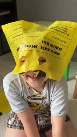 A young child with a bright yellow plastic bag over their head, with two eyeholes cut out, smiles at the camera. The bag is decorated with text in Cyrillic and has a logo that appears to be for a women's clothing store. The child is wearing a light gray t-shirt with a cartoon character graphic and a beaded necklace. They are sitting on a light-colored floor, possibly a carpet or playmat. In the background, a green object and part of a wooden chair are visible, along with a bookshelf. The lighting suggests it is daytime. The text on the bag reads "Магазин одежды" (Clothing store) and includes a phone number and what appears to be an address or store information. The child's expression is joyful and playful. The location is identified as Boraldai, Kazakhstan.
