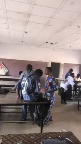 Several people are inside a classroom. There are tables and chairs, and some people are standing around the tables. The room is plain, with a white ceiling and brown walls.  The lighting appears to be coming from windows along one wall. One person is wearing a patterned blue and orange outfit, and another wears a blue shirt with writing on it. Some of the people have backpacks.