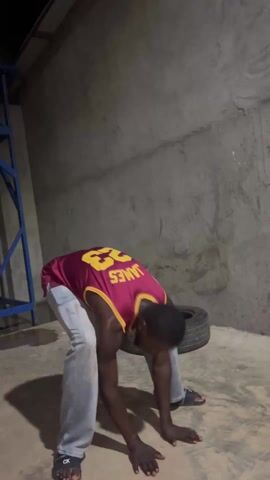 A man in a red and gold basketball jersey and light-colored jeans dances in a cement room. He wears black sandals. There is a tire behind him, and a metal table against the wall. The room is lit dimly.