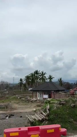 The video shows a panning view of a rural area with some construction or demolition happening. In the foreground, there is a red construction barrier with yellow and black hazard stripes and some graffiti. Beyond the barrier, there are piles of logs, green grass, and dirt. A small building with a dark roof and white walls is visible, surrounded by some unfinished brickwork. Further back, there are more buildings with corrugated metal roofs, some of which appear damaged or unfinished. Palm trees are scattered throughout the landscape, and there are power lines and utility poles visible. The sky is overcast with clouds. The overall impression is one of a developing or rebuilding area.