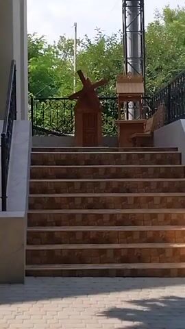 The video shows a set of outdoor stairs leading up to a landing with several decorative objects. In the center of the landing is a small, wooden windmill-like structure with a door. To its right is a wooden well with a pitched roof, and a bench attached to its side. A metal railing runs along the left side of the stairs and continues along the landing. Behind the landing is a black metal fence, and beyond that, lush green trees and foliage. A tall, grey, cylindrical structure, possibly a chimney or industrial pipe, is visible on the far right, extending upwards. The sky is overcast. The stairs appear to be tiled with a mosaic pattern. The ground in front of the stairs is paved with light-colored interlocking bricks. The overall scene appears to be an outdoor recreational or decorative area.
