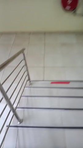 A person in brown shoes and camo pants walks down a set of stairs. The stairs have white tile steps with a gray metal rail along the side. There is a red strip of tape on one of the steps. The wall is off-white and tiled. The camera follows the person's feet as they descend.